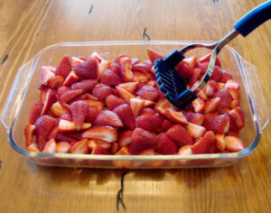 Fresh strawberries in 9x13 dish getting mashed with potato masher.