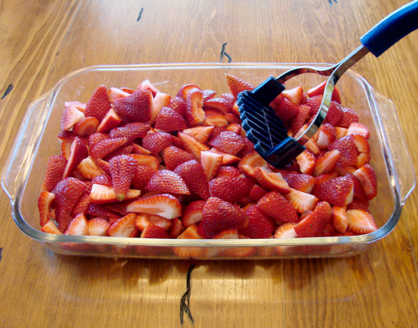 Fresh strawberries in 9x13 dish getting mashed with potato masher.