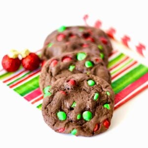 Three chocolate M&M Christmas cookies on napkin with ornaments to the side.