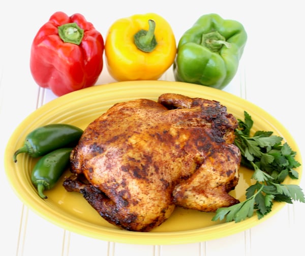 Cooked whole chicken on platter with three bell peppers in the background.