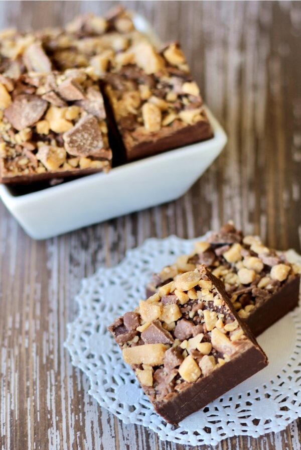Two pieces of chocolate 3 ingredient fudge on doily, with more fudge pieces in background.