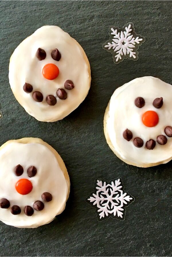 Three frosted snowman cookies decorated with chocolate chip eyes and mouths and m&m noses.