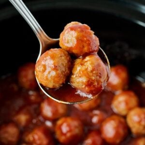 Three cocktail meatballs on spoon with crockpot full of meatballs in background.