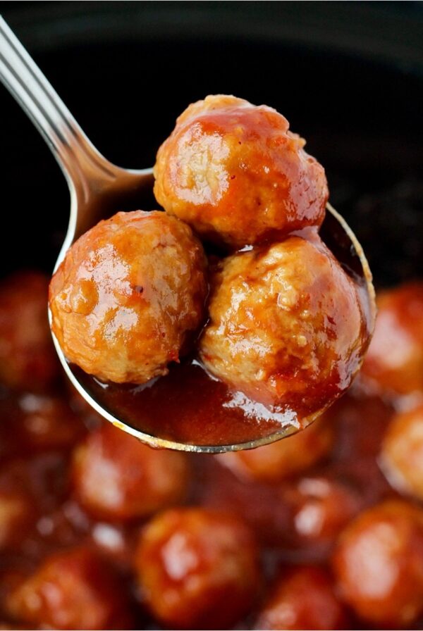 Three cocktail meatballs on spoon with crockpot full of meatballs in background.