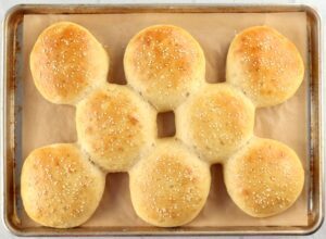 Cooked hamburger buns with sesame seeds on a lined baking sheet.