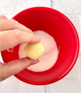 Person holding a ball of dough over a red bowl of sugar.