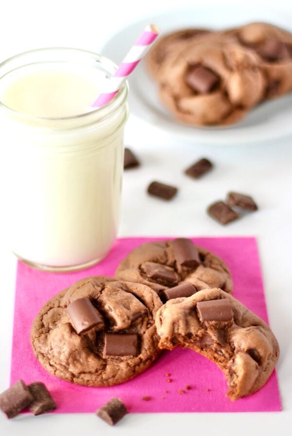 Three chewy chocolate chunk cookies on napkin with chocolate chunks and glass of milk to the side.