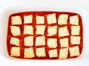 Casserole dish with marinara sauce and rows of frozen ravioli.