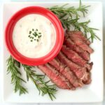 Horseradish sauce in red dish on plate with sliced steak and rosemary sprigs on the side.