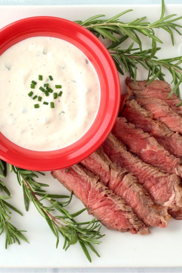 Horseradish sauce in red dish on plate with sliced steak and rosemary sprigs on the side.