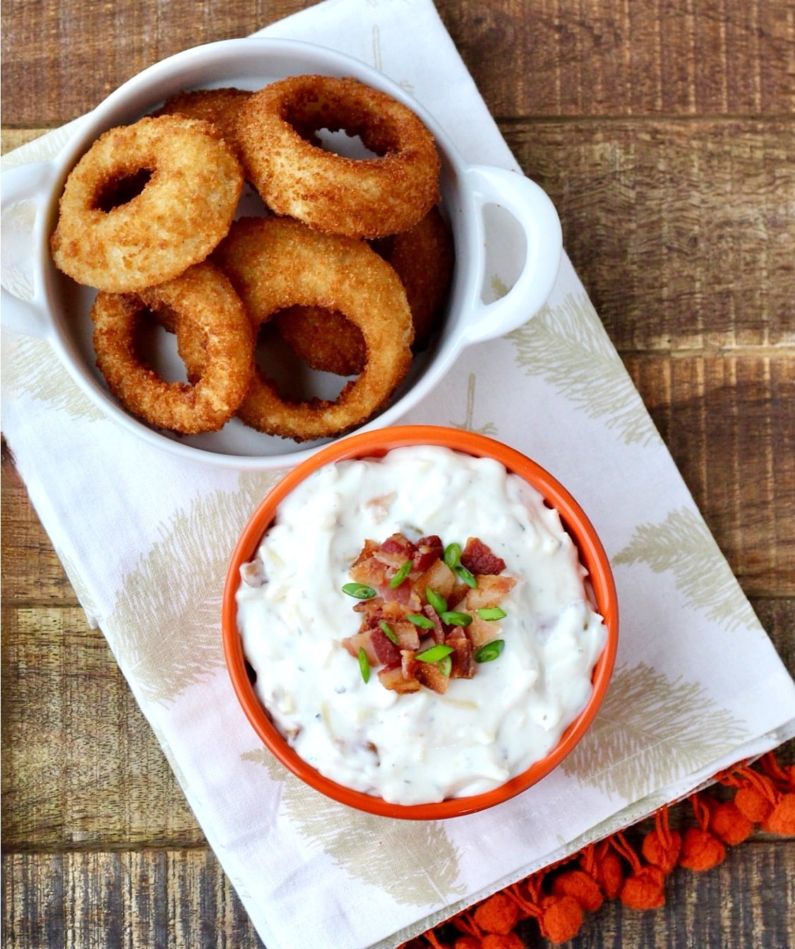 Homemade ranch dip in bowl with bacon and chives on top, and onion rings to the side.