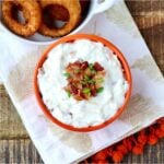 Ranch dip in bowl with bacon and chives on top, and onion rings to the side.