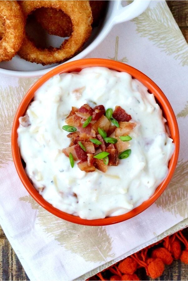 Ranch dip in bowl with bacon and chives on top, and onion rings to the side.
