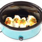 Four pieces of chicken in crockpot with ranch sauce, bacon, cheddar, and green onions on top.