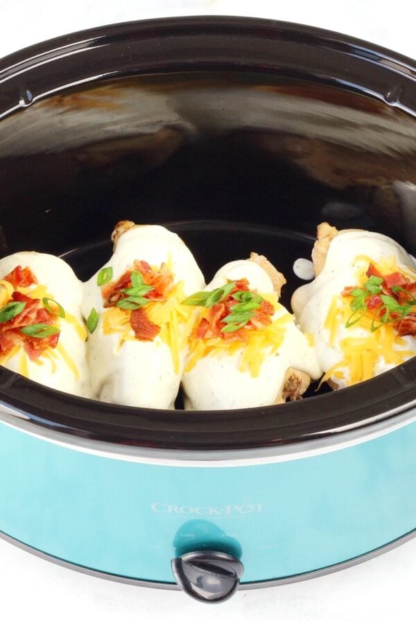 Four pieces of chicken in crockpot with ranch sauce, bacon, cheddar, and green onions on top.