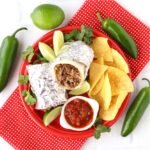 Beef burrito on plate with chips, salsa, lime, cilantro, and jalapeño peppers to the side.
