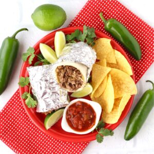 Beef burrito on plate with chips, salsa, lime, cilantro, and jalape&ntilde;o peppers to the side.