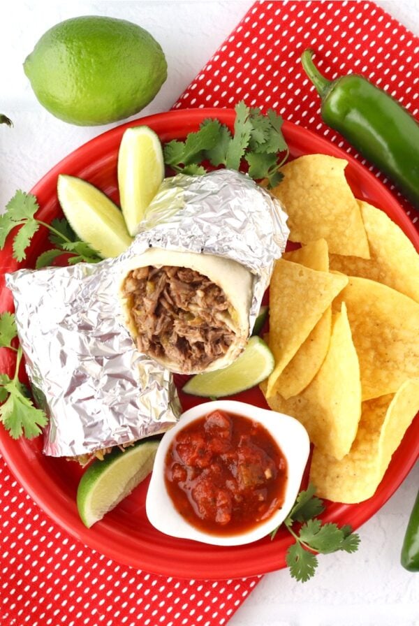 Beef burrito on plate with chips, salsa, lime, cilantro, and jalapeño peppers to the side.