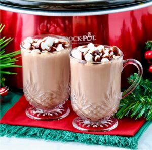 Two mugs of hot chocolate with marshmallows in front of crockpot.