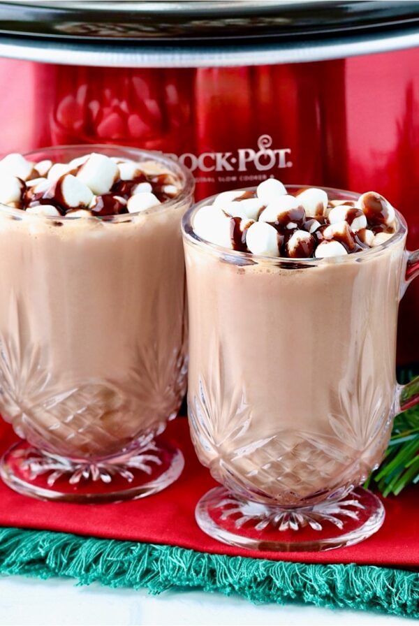 Two mugs of hot chocolate with marshmallows in front of crockpot.
