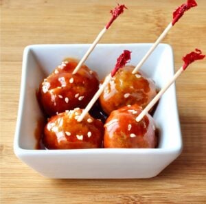 Four meatballs in small dish with honey garlic sauce, garnished with sesame seeds and toothpicks inserted.