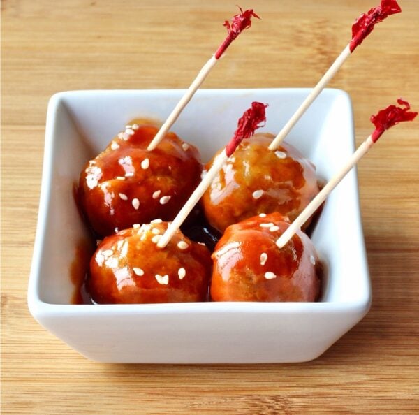Four meatballs in small dish with honey garlic sauce, garnished with sesame seeds and toothpicks inserted.