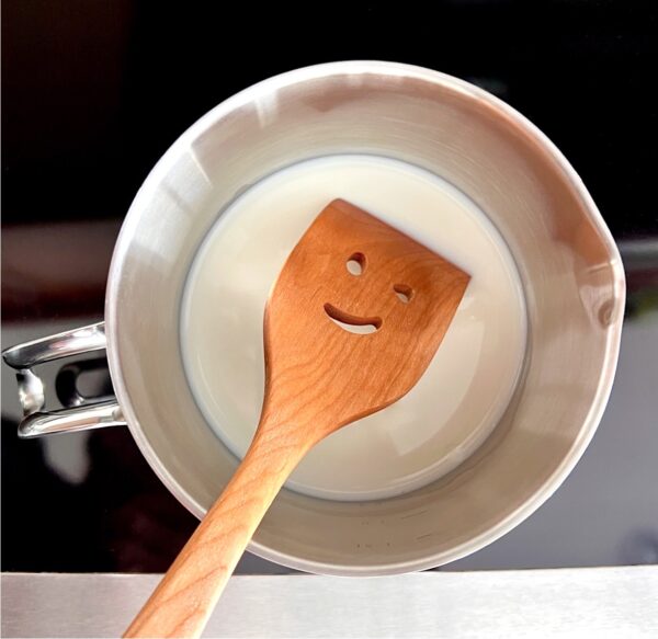 Milk for Peppermint Mocha warming on stove with smiling wooden spoon.
