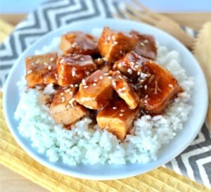 Cubed orange chicken on plate with rice, garnished with sesame seeds.