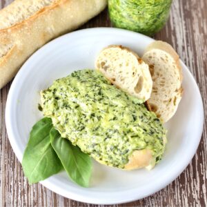 Piece of pesto chicken on plate with slices of bread and basil leaves to the side.
