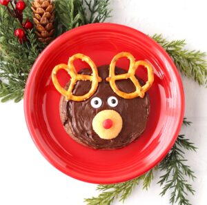 Frosted chocolate Rudolph reindeer cookie with pretzel ears, candy eyes, and Nilla wafer nose on plate.