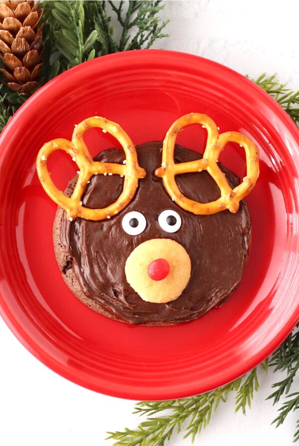 Frosted chocolate Rudolph reindeer cookie with pretzel ears, candy eyes, and Nilla wafer nose on plate.