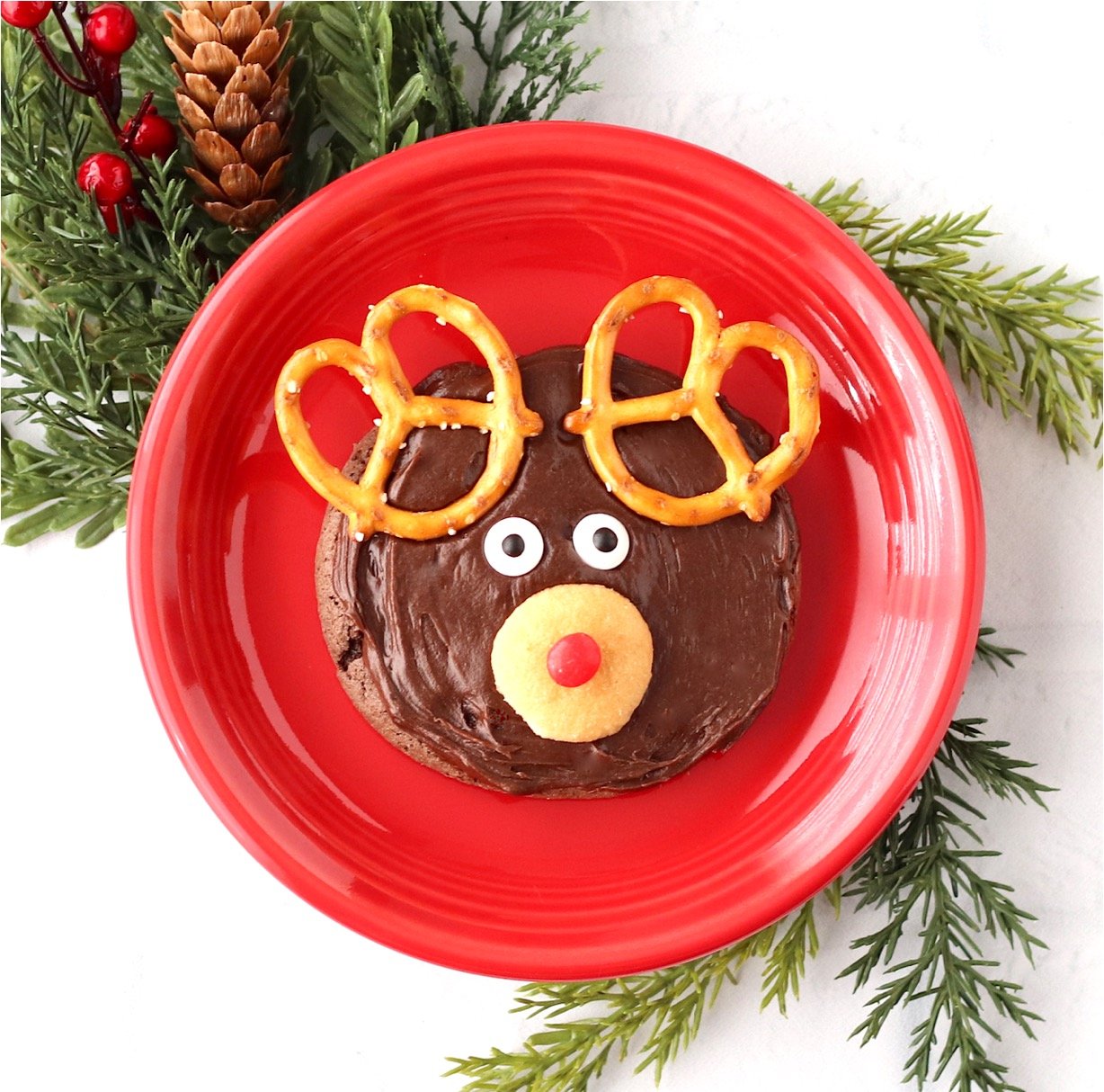 Frosted chocolate Rudolph reindeer cookie with pretzel ears, candy eyes, and Nilla wafer nose on plate.