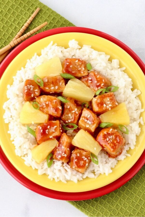 Sweet and sour pork with pineapples, green onions and sesame seeds on plate with rice.