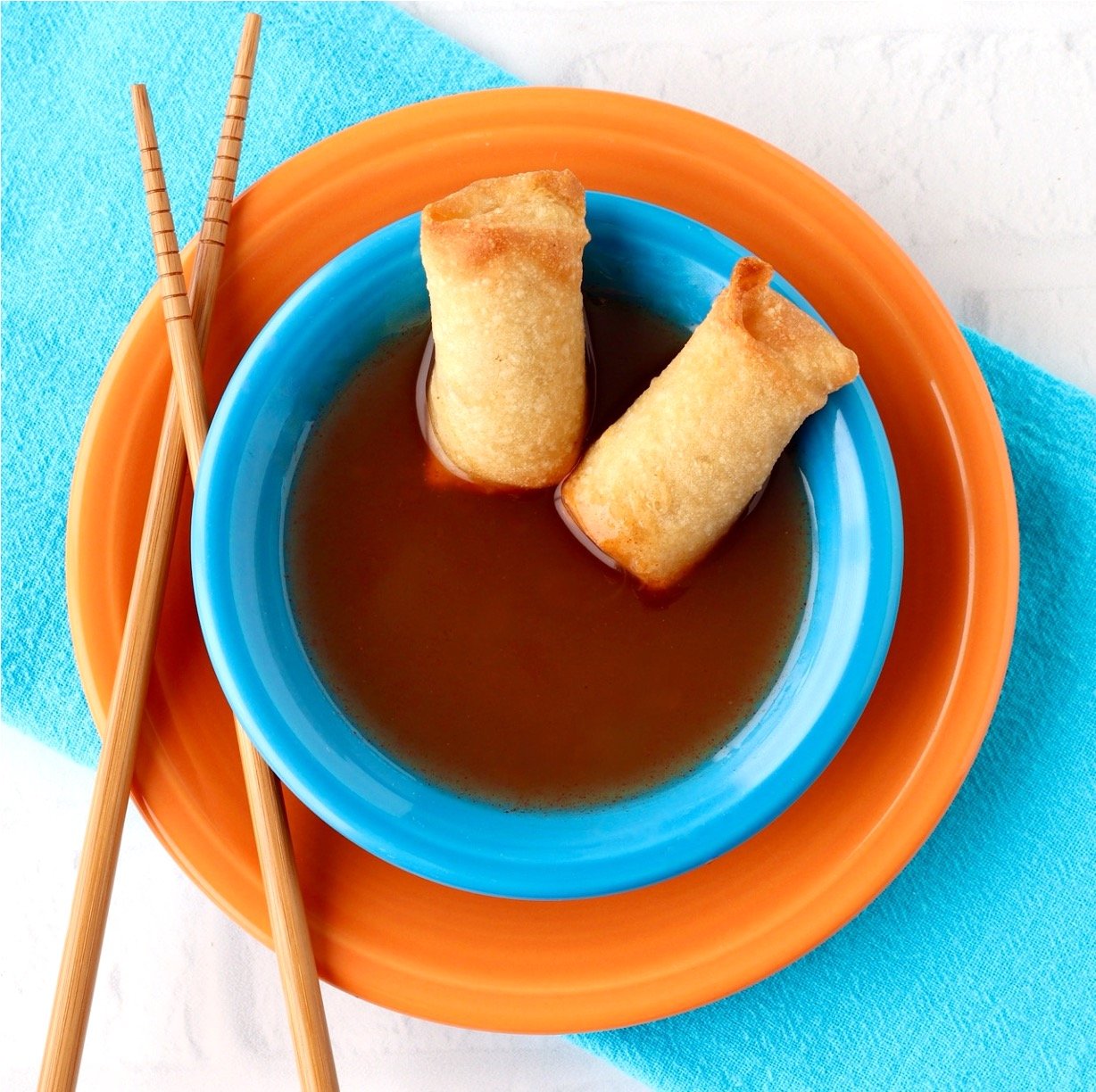 Two egg rolls in bowl with sweet and sour sauce, and chopsticks to the side.