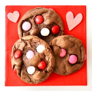 Three chocolate M&M Valentine's Day Cookies on napkin with hearts to the side.