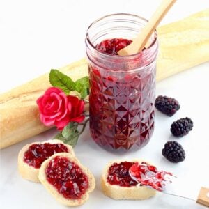 Homemade blackberry jam in mason jar with spoon, and bread slices with jam on top to the side.