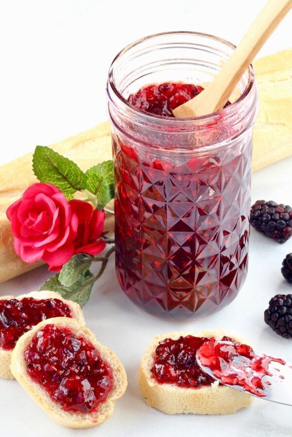 Homemade blackberry jam in mason jar with spoon, and bread slices with jam on top to the side.