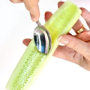 Hands holding peeled cucumber while spoon is removing seeds.