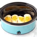 Four pieces of chicken in crock pot with creamy garlic sauce, cheddar, and green onions on top.