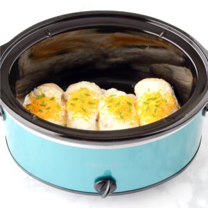 Four pieces of chicken in crock pot with creamy garlic sauce, cheddar, and green onions on top.