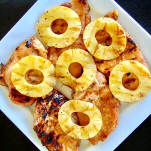 Five pieces of grilled teriyaki chicken on plate topped with grilled pineapple rings.