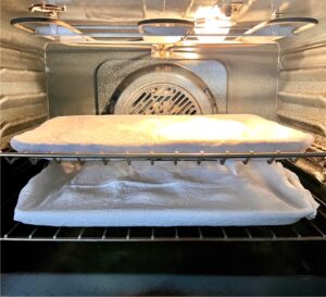 Two trays of slider buns covered with flour sack cloth proofing in oven with light on.