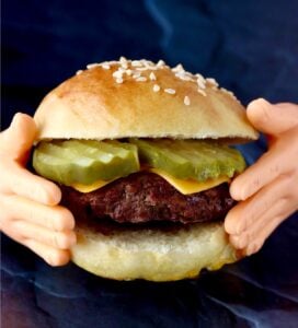 Tiny hands holding small burger with cheese and pickles on mini slider bun.