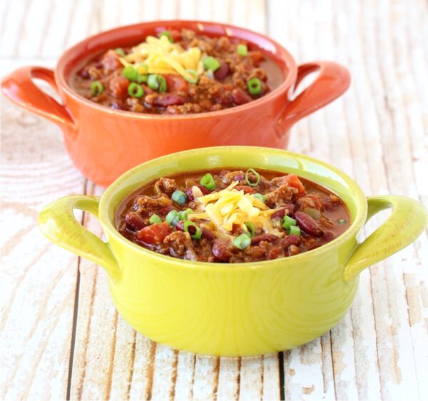 Two bowls of ground beef chili with shredded cheddar cheese and green onions on top.