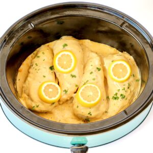 Four pieces of chicken in crockpot topped with lemon garlic sauce, lemon slices and parsley.