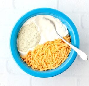 Sour cream, ranch seasoning, and shredded cheddar in mixing bowl with spoon for ranch dip.