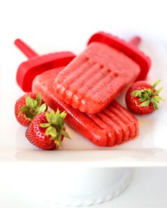 Two strawberry popsicles on plate, surrounded by fresh strawberries.