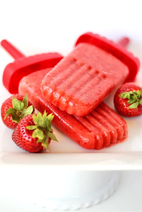 Two strawberry popsicles on plate, surrounded by fresh strawberries.