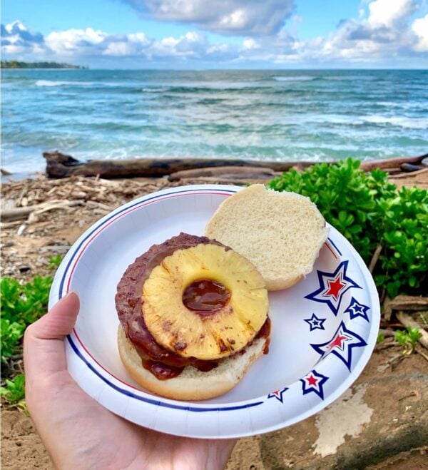 Teriyaki burger with grilled pineapple ring on top on paper plate at the beach.