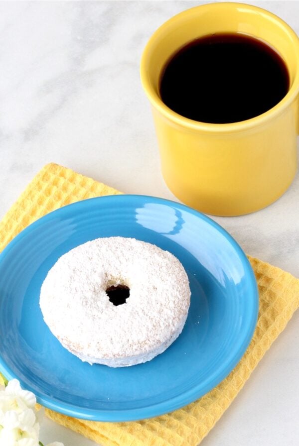 Powdered sugar donut on plate with cup of coffee to the side.